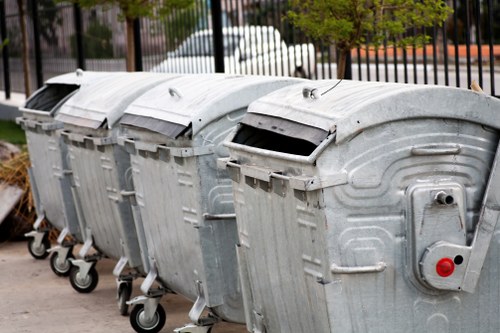 A row of four large, metallic rubbish bins with curved lids, positioned on a paved surface likely within a residential or commercial area in Finchley. The bins are made of galvanized steel with a textured, slightly reflective surface, showing some signs of wear and scratches. Each bin has a black handle on the front edge of the lid and is mounted on black rubber wheels for mobility, with the front wheels visible in the foreground. The bins' lids are closed and have small central protrusions, and the bins are lined up side by side, creating a uniform appearance. The background features a black metal fence and blurred trees, suggesting an outdoor setting with natural light. These bins are commonly used for waste collection in local rubbish removal services, such as those offered by Garden Clearance Finchley, supporting sustainable waste management in the area.