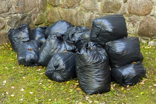 A collection of ten large black plastic rubbish bags piled on a grassy area near a stone wall, with some bags leaning against the wall and others stacked on top of each other. The bags are tightly sealed and appear to contain garden or general waste, with a slightly wrinkled texture and a glossy finish. The grass surrounding the bags is patchy with small fallen leaves, indicating an outdoor setting likely in a residential garden or driveway in Finchley. In the background, the stone wall provides a rustic, natural backdrop, framing the scene. The image suggests rubbish removal or clearance activity, typical of services offered by Garden Clearance Finchley in the local area, with the bags ready for collection or disposal at a nearby waste facility.