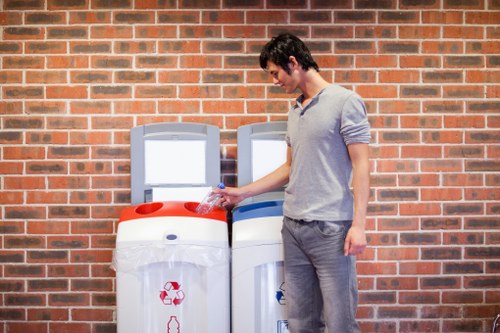 A young man with dark hair, wearing a light grey long-sleeved shirt and grey trousers, stands outdoors against a red brick wall. He is positioned beside two white recycling bins with contrasting blue and red lids, and he is seen placing a clear plastic bottle into the bin with the red lid. The bins are placed on a paved surface, and the scene suggests a residential or community area likely within the Finchley region, related to waste management or rubbish collection services. The lighting is natural, indicating daytime, and the environment appears clean and orderly, with no other objects or debris visible in the immediate vicinity. This setting exemplifies rubbish disposal practices within a UK urban context, supporting services like those offered by Garden Clearance Finchley for rubbish removal and waste disposal.
