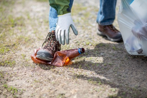 A person wearing light-colored gloves and brown leather boots is shown picking up a discarded brown glass bottle from a patchy outdoor ground surface covered with dirt and small grass patches. The individual is dressed in green trousers, and nearby, a grey plastic rubbish bag is partially visible to the right. The scene suggests an outdoor environment, possibly a garden or area within Finchley, with a focus on waste collection or rubbish removal activities typical of services provided by Garden Clearance Finchley. The background features a gravel or dirt pathway with some green grass, indicating a typical setting for garden and rubbish clearance tasks. The overall atmosphere is neutral, emphasizing the action of waste removal with detailed attention to the texture of the bottle, the ground surface, and the clothing on the person, supporting the context of professional rubbish clearance services in the local area.
