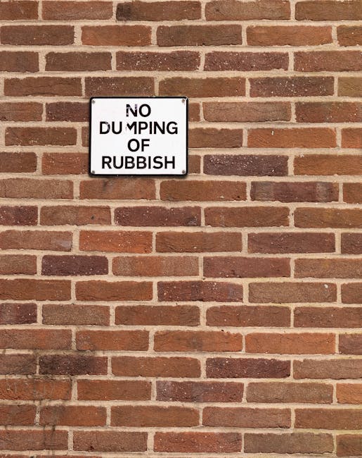 A pair of large black plastic rubbish bags and a cardboard box filled with waste materials are placed on the pavement in front of a dark metal fence and dense foliage. The bags are crumpled and appear