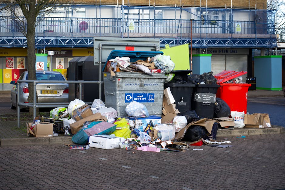 A small, light blue utility truck is parked on the side of a road, loaded with an extensive amount of waste materials. The cargo area is filled with flattened cardboard boxes, black plastic garbage ba